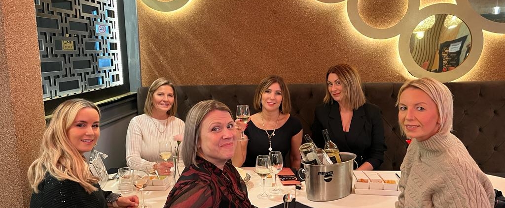6 Women sat around a table looking at the camera smiling