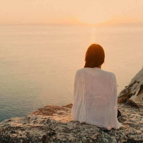 a woman sat on the edge of a cliff, overlooking a sunset by the sea