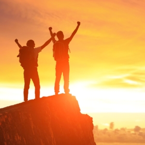 Two male silhouettes standing on the edge of a rock in the sunset