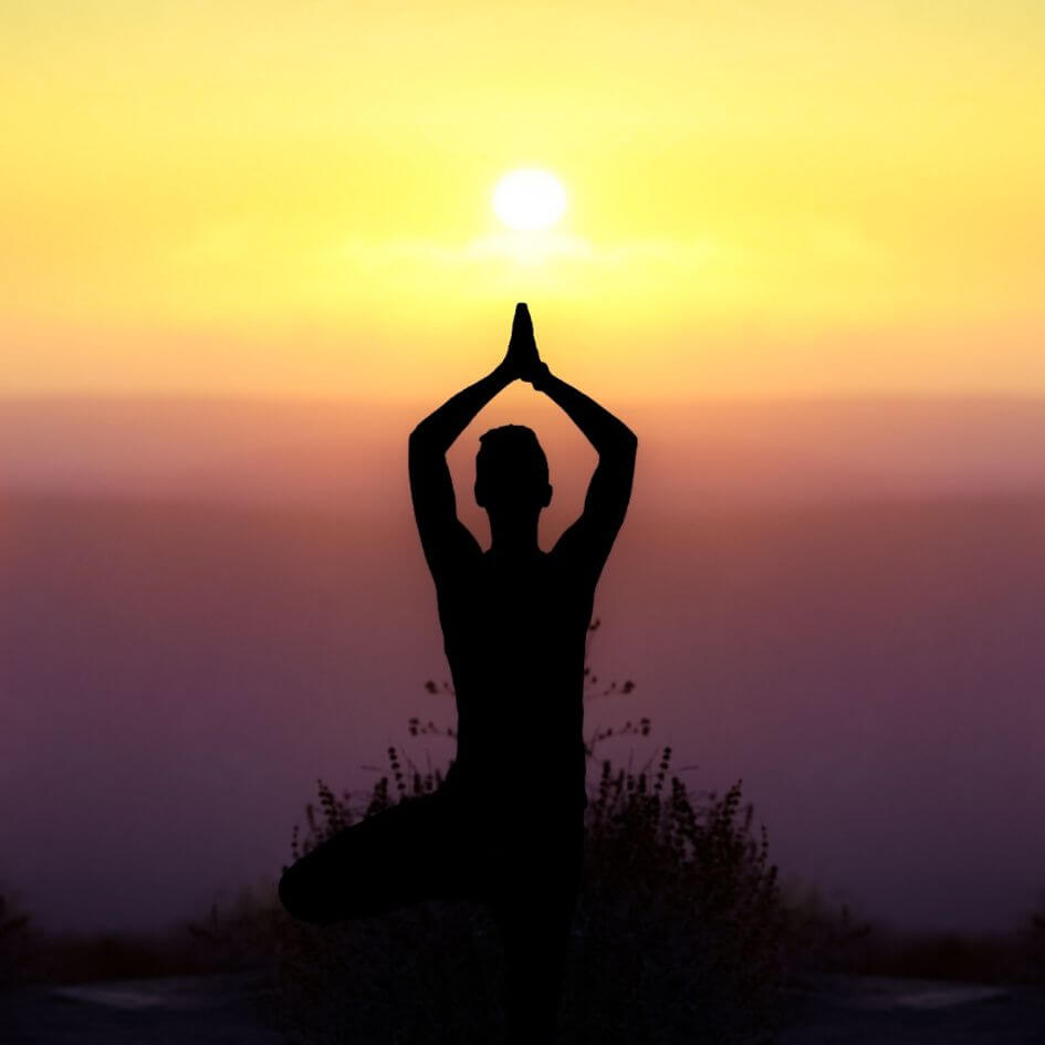 a silhouette of a woman in a yoga pose on a click edge with the sun setting in the distance