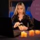 Women holding tarot cards surrounded by candles whilst at a computer